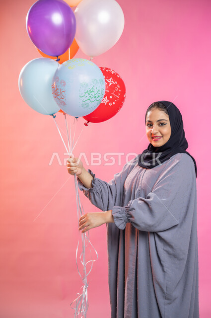 التسلية والترفيه وقضاء أوقات ممتعة، بورتريه لامرأة عربية سعودية خليجية محجبة ترتدي العباءة و  تحمل بيديها مجموعة بوالين ملونة ، إيماءات تدل على الفرح والسرور بالعيد السعيد , مناسبات واعياد إسلامية. 