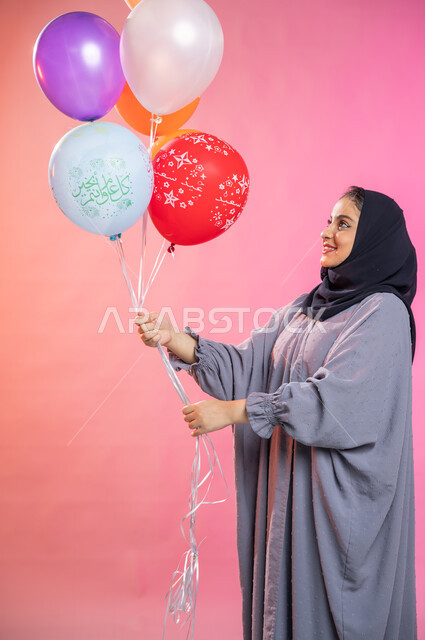 التسلية والترفيه وقضاء أوقات ممتعة، بورتريه لامرأة عربية سعودية خليجية محجبة ترتدي العباءة و  تحمل بيديها مجموعة بوالين ملونة ، إيماءات تدل على الفرح والسرور بالعيد السعيد , مناسبات واعياد إسلامية.