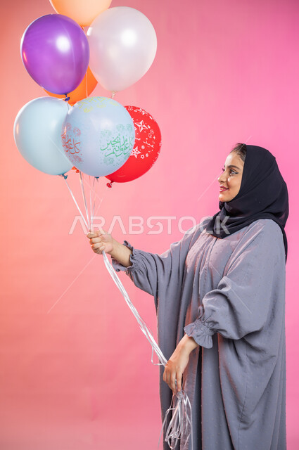 إيماءات تدل على الفرح والسرور بالعيد السعيد ,التسلية والترفيه وقضاء أوقات ممتعة، بورتريه لامرأة عربية سعودية خليجية محجبة ترتدي العباءة و  تحمل بيديها مجموعة بالونات بألوان زاهية ، مناسبات واعياد إسلامية، خلفية ملونة. 