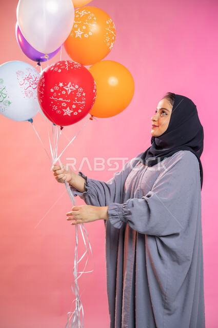 التسلية والترفيه وقضاء أوقات ممتعة، بورتريه لامرأة عربية سعودية خليجية محجبة ترتدي العباءة و  تحمل بيديها مجموعة بالونات بألوان زاهية ، مناسبات وأعياد إسلامية، خلفية ملونة.