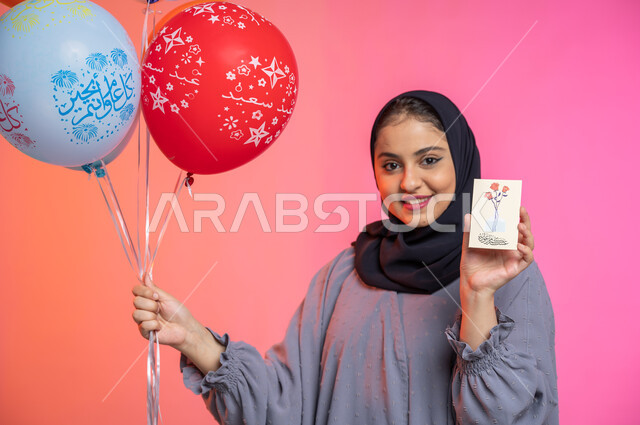 A congratulatory card for the advent of Eid Al-Adha, a portrait of a veiled Saudi Arab Gulf woman wearing a gown and holding a greeting card in her hand and a group of bawali, happy Islamic holidays and occasions, facial gestures indicating happiness and 