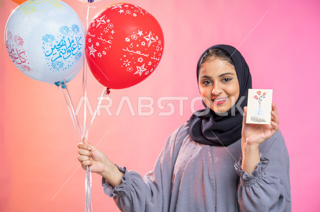 بطاقة تهنئة بقدوم عيد الأضحى المبارك ، بورتريه لسيدة عربية خليجية سعودية محجبة ترتدي عباءة وتحمل بيدها بطاقة المعايدة ومجموعة بالونات ، أعياد ومناسبات إسلامية سعيدة، إيماءات وجه تدل على السعادة والفرح، خلفية ملونة.