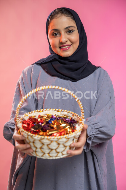 إيماءات وجه تدل على البشاشة والترحيب بالضيوف, بورتريه لامرأة عربية سعودية خليجية محجبة ترتدي العباءة تحمل بيدها سلة من القش مليئة بالحلوى و السكاكر , حسن الضيافة في الأعياد و المناسبات , خلفية ملونة.