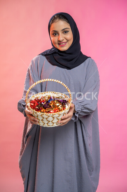 إيماءات وجه تدل على البشاشة والترحيب بالضيوف, بورتريه لامرأة عربية سعودية خليجية محجبة ترتدي العباءة تحمل بيدها سلة من القش مليئة بالحلوى و السكاكر , حسن الضيافة في الأعياد و المناسبات , خلفية ملونة.