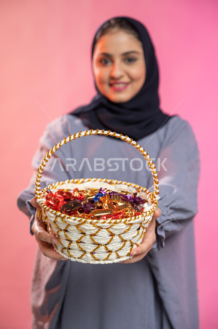 حسن الضيافة في الأعياد و المناسبات ,إيماءات وجه تدل على البشاشة والترحيب بالضيوف, بورتريه لامرأة عربية سعودية خليجية محجبة ترتدي العباءة تحمل بيدها سلة من القش مليئة بالحلوى و السكاكر , خلفية ملونة.