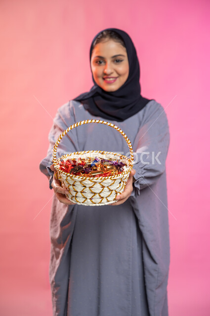 حسن الضيافة في الأعياد و المناسبات ,إيماءات وجه تدل على البشاشة والترحيب بالضيوف, بورتريه لامرأة عربية سعودية خليجية محجبة ترتدي العباءة تحمل بيدها سلة من القش مليئة بالحلوى و السكاكر , خلفية ملونة.