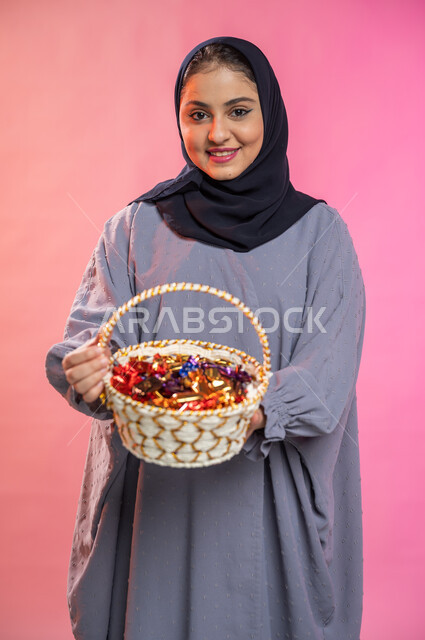 حسن الضيافة في الأعياد و المناسبات ,إيماءات وجه تدل على البشاشة والترحيب بالضيوف, بورتريه لامرأة عربية سعودية خليجية محجبة ترتدي العباءة تحمل بيدها سلة من القش مليئة بالحلوى و السكاكر , خلفية ملونة.