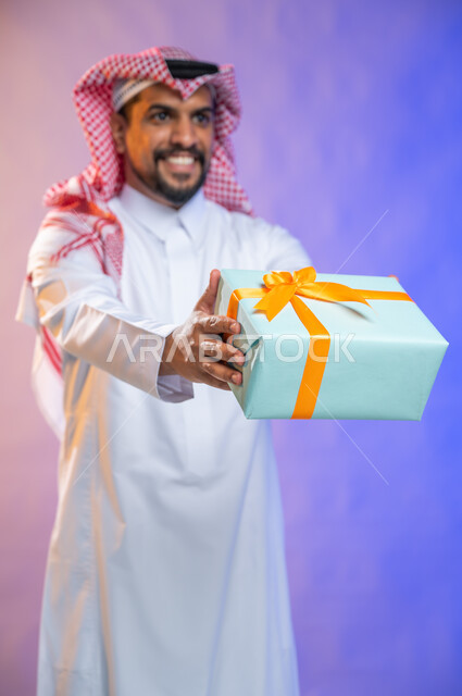 أعياد ومناسبات ومفاجآت سعيدة, إيماءات وجه ويدين تدل على الفرح والسعادة بالهدايا و التهنئات، بورتريه لشاب عربي سعودي خليجي يرتدي الثوب و الشماغ السعودي التقليدي يحمل بيده علبة هدايا، خلفية ملونة.