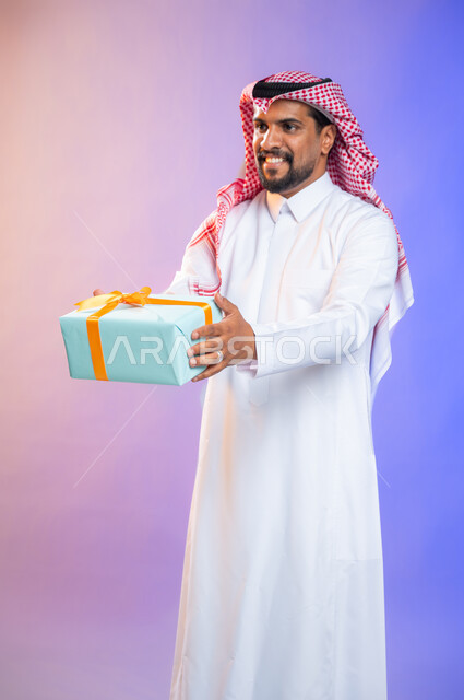 أعياد ومناسبات ومفاجآت سعيدة, إيماءات وجه ويدين تدل على الفرح والسعادة بالهدايا و التهنئات، بورتريه لشاب عربي سعودي خليجي يرتدي الثوب و الشماغ السعودي التقليدي يحمل بيده علبة هدايا، خلفية ملونة.