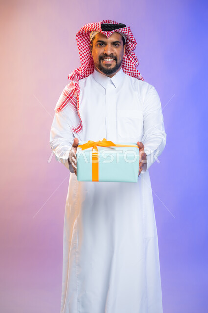 أعياد ومناسبات ومفاجآت سعيدة, إيماءات وجه ويدين تدل على الفرح والسعادة بالهدايا و التهنئات، بورتريه لشاب عربي سعودي خليجي يرتدي الثوب و الشماغ السعودي التقليدي يحمل بيده علبة هدايا، خلفية ملونة.