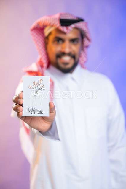 بهجه وفرحة استقبال العيد، بورتريه لرجل خليجي سعودي عربي يرتدي الثوب السعودي التقليدي والشماغ يمسك بيده بطاقة معايدة، الاحتفال بالأعياد والمناسبات الإسلامية السعيدة، خلفية ملونة، بطاقات تهنئة بعيد الأضحى، ايماءات تدل على الفرح والسرور 
