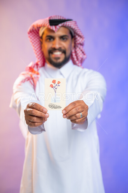 بهجه وفرحة استقبال العيد، بورتريه لرجل خليجي سعودي عربي يرتدي الثوب السعودي التقليدي والشماغ يمسك بيده بطاقة معايدة، الاحتفال بالأعياد والمناسبات الإسلامية السعيدة، خلفية ملونة، بطاقات تهنئة بعيد الأضحى، ايماءات تدل على الفرح والسرور 