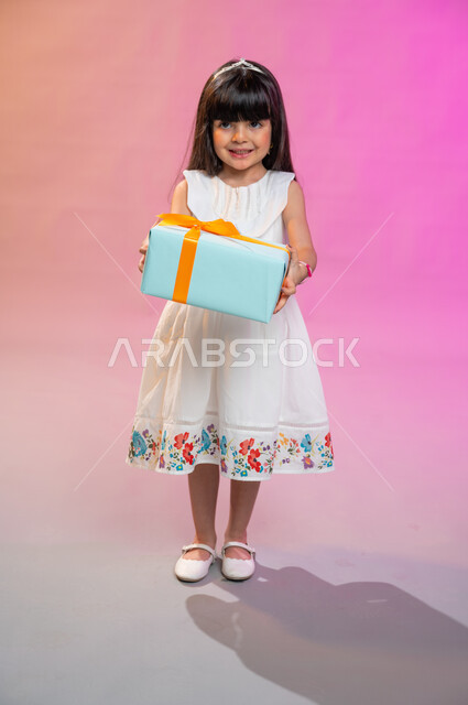 The joy of children with the gift of Eid, holidays, occasions and happy surprises, face and hand gestures indicating joy and happiness with gifts and congratulations, a portrait of a Saudi Arabian Gulf girl wearing a beautiful dress holding a gift box in 