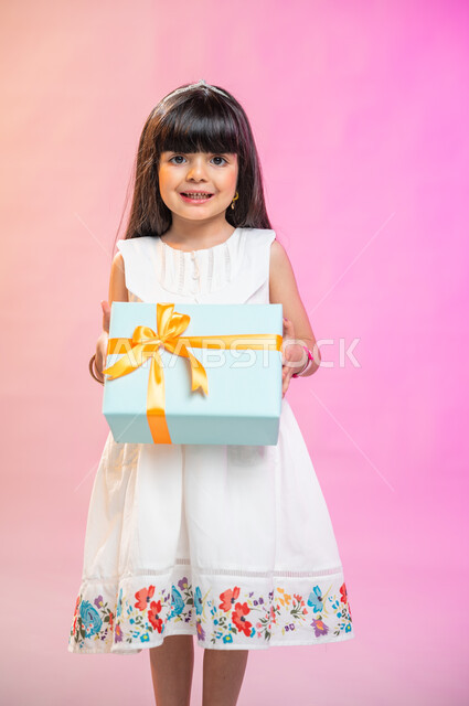 أعياد ومناسبات ومفاجآت سعيدة, فرحة الأطفال بهدية العيد، إيماءات وجه ويدين تدل على الفرح والسعادة بالهدايا و التهنئات، بورتريه لطفلة عربية سعودية خليجية ترتدي فستان جميل تحمل بيديها علبة هدايا، خلفية ملونة. 