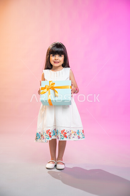 أعياد ومناسبات ومفاجآت سعيدة, فرحة الأطفال بهدية العيد، إيماءات وجه ويدين تدل على الفرح والسعادة بالهدايا و التهنئات، بورتريه لطفلة عربية سعودية خليجية ترتدي فستان جميل تحمل بيديها علبة هدايا، خلفية ملونة، صورة الجسم بالطول الكامل. 