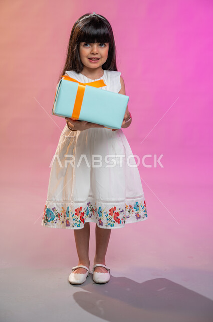 أعياد ومناسبات ومفاجآت سعيدة, فرحة الأطفال بهدية العيد، إيماءات وجه ويدين تدل على الفرح والسعادة بالهدايا و التهنئات، بورتريه لطفلة عربية سعودية خليجية ترتدي فستان جميل تحمل بيديها علبة هدايا، خلفية ملونة، صورة الجسم بالطول الكامل. 