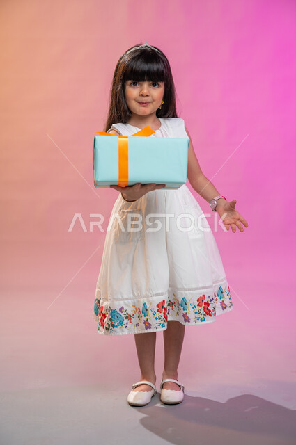 أعياد ومناسبات ومفاجآت سعيدة, فرحة الأطفال بهدية العيد، إيماءات وجه ويدين تدل على الفرح والسعادة بالهدايا و التهنئات، بورتريه لطفلة عربية سعودية خليجية ترتدي فستان جميل تحمل بيديها علبة هدايا، خلفية ملونة، صورة الجسم بالطول الكامل. 