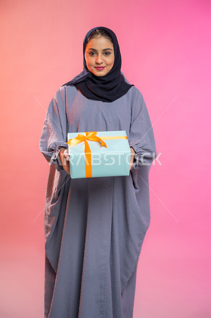 أعياد ومناسبات ومفاجآت سعيدة, إيماءات وجه تدل على الفرح والسعادة بالهدايا و التهنئات، بورتريه لامرأة عربية سعودية خليجية محجبة ترتدي عباءة تحمل بيديها علبة هدية مغلفة، خلفية ملونة. 