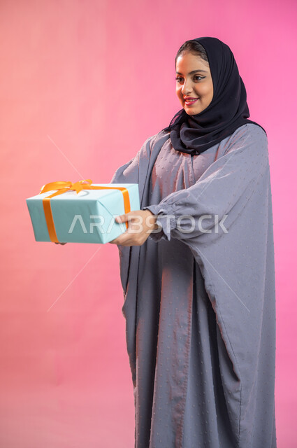 Holidays, occasions and happy surprises, facial gestures indicating joy and happiness with gifts and congratulations, a portrait of a veiled Saudi Arab Gulf woman wearing an abaya holding a wrapped gift box in her hands, colored background.