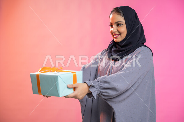 أعياد ومناسبات ومفاجآت سعيدة, إيماءات وجه تدل على الفرح والسعادة بالهدايا و التهنئات، بورتريه لامرأة عربية سعودية خليجية محجبة ترتدي عباءة تحمل بيديها علبة هدية مغلفة، خلفية ملونة. 