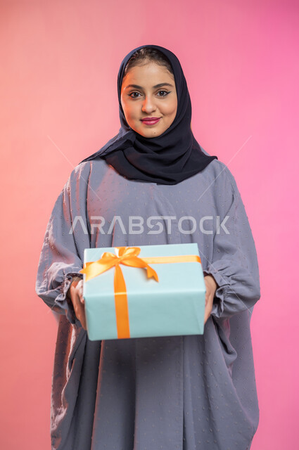 أعياد ومناسبات ومفاجآت سعيدة, إيماءات وجه تدل على الفرح والسعادة بالهدايا و التهنئات، بورتريه لامرأة عربية سعودية خليجية محجبة ترتدي عباءة تحمل بيديها علبة هدية مغلفة، خلفية ملونة.