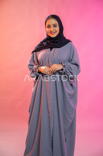 ايماءات وجه و ابتسامة سعادة و سرور, بورتريه لامرأة عربية سعودية خليجية محجبة ترتدي عباءة تقف باستقامة وتنظر نحو الكاميرا,  خلفية ملونة.