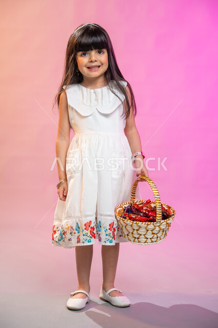صورة الجسم بالطول الكامل، بهجه وفرحة الأطفال بالعيد السعيد، بورتريه فتاة عربية خليجية سعودية ترتدي فستان أبيض تحمل سلة قش مليئة بالسكاكر،  تقديم حلوى والقرقيعان ، عادات العيد في المجتمع السعودي الخليجي، أعياد ومناسبات إسلامية سعيدة ، خلفية ملونة