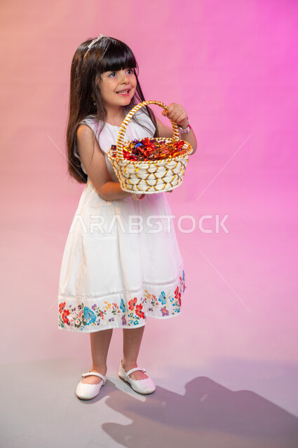 صورة الجسم بالطول الكامل، بهجه وفرحة الأطفال بالعيد السعيد، بورتريه فتاة عربية خليجية سعودية ترتدي فستان أبيض تحمل سلة قش مليئة بالسكاكر،  تقديم حلوى والقرقيعان ، عادات العيد في المجتمع السعودي الخليجي، أعياد ومناسبات إسلامية سعيدة ، خلفية ملونة