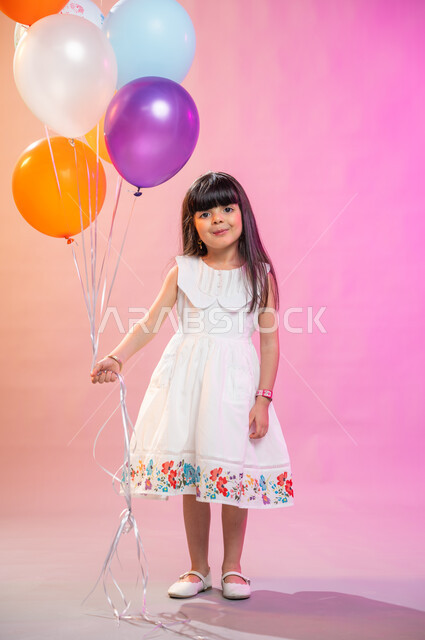 صورة الجسم بالطول الكامل، بهجة وفرحة الأطفال بالعيد السعيد، بورتريه فتاة عربية خليجية سعودية ترتدي فستان أبيض تمسك مجموعة بالونات، عادات الأعياد في المجتمع السعودي الخليجي، التحضير والتجهيز للاحتفال بالأعياد والمناسبات الإسلامية السعيدة ، خلفية ملونة