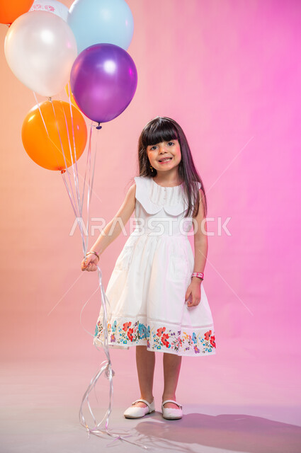 صورة الجسم بالطول الكامل، بهجة وفرحة الأطفال بالعيد السعيد، بورتريه فتاة عربية خليجية سعودية ترتدي فستان أبيض تمسك مجموعة بالونات، عادات الأعياد في المجتمع السعودي الخليجي، التحضير والتجهيز للاحتفال بالأعياد والمناسبات الإسلامية السعيدة ، خلفية ملونة