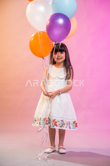 صورة الجسم بالطول الكامل، بهجة وفرحة الأطفال بالعيد السعيد، بورتريه فتاة عربية خليجية سعودية ترتدي فستان أبيض تمسك مجموعة بالونات، عادات الأعياد في المجتمع السعودي الخليجي، التحضير والتجهيز للاحتفال بالأعياد والمناسبات الإسلامية السعيدة ، خلفية ملونة