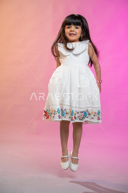 إيماءات تدل على الفرح والحماس والبهجة، بورتريه لطفلة عربية سعودية خليجية ترتدي فستان جميل تقفز فرحا وسعادة، صورة الجسم بالطول الكامل، خلفية ملونة