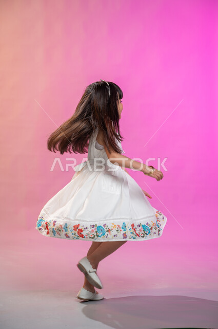 إيماءات تدل على الفرح والحماس والبهجة، بورتريه لطفلة عربية سعودية خليجية ترتدي فستان جميل تقفز فرحا وسعادة، صورة الجسم بالطول الكامل، خلفية ملونة