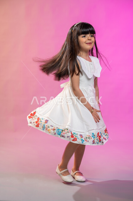 فرحة الأطفال بثياب العيد الجديدة ، إيماءات تدل على الفرح والبهجة، بورتريه لطفلة عربية سعودية خليجية ترتدي فستان جميل تقفز فرحا وسعادة، صورة الجسم بالطول الكامل، خلفية ملونة