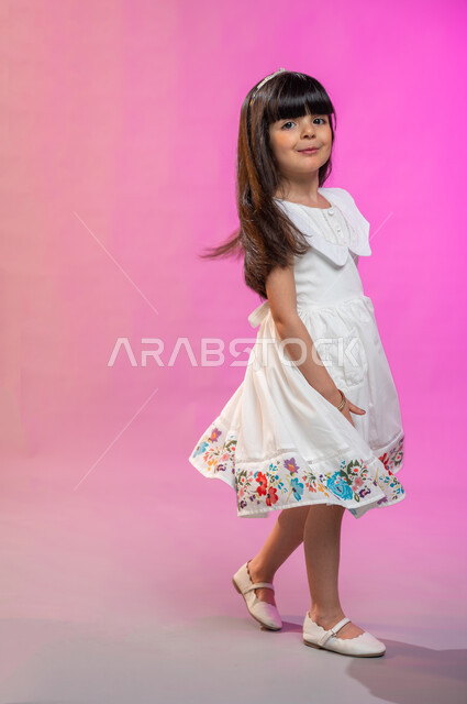 فرحة الأطفال بثياب العيد الجديدة ، إيماءات تدل على الفرح والبهجة، بورتريه لطفلة عربية سعودية خليجية ترتدي فستان جميل تقفز فرحا وسعادة، صورة الجسم بالطول الكامل، خلفية ملونة