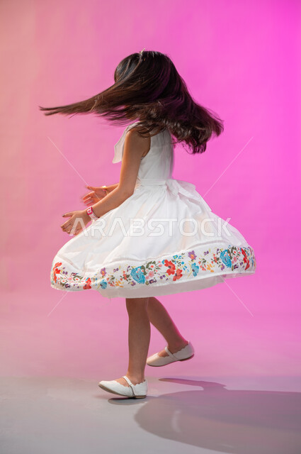 فرحة الأطفال بثياب العيد الجديدة ، إيماءات تدل على الفرح والبهجة، بورتريه لطفلة عربية سعودية خليجية ترتدي فستان جميل تقفز فرحا وسعادة، صورة الجسم بالطول الكامل، خلفية ملونة