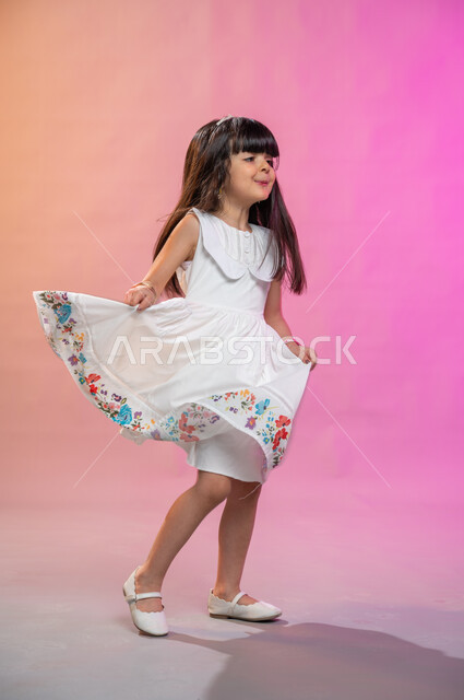 فرحة الأطفال بثياب العيد الجديدة ، إيماءات تدل على الفرح والبهجة، بورتريه لطفلة عربية سعودية خليجية ترتدي فستان جميل تقفز فرحا وسعادة، صورة الجسم بالطول الكامل، خلفية ملونة