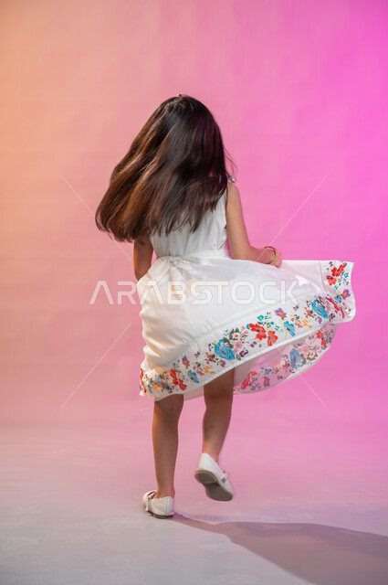 فرحة الأطفال بثياب العيد الجديدة ، إيماءات تدل على الفرح والبهجة، بورتريه لطفلة عربية سعودية خليجية ترتدي فستان جميل تقفز فرحا وسعادة، صورة الجسم بالطول الكامل، خلفية ملونة