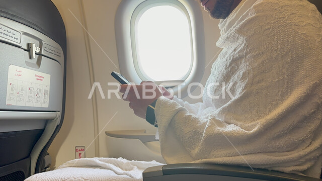 استخدام الهاتف المحمول, صورة مقربة لرجل عربي خليجي سعودي يجلس في مقصورة الطائرة يرتدي لباس الاحرام , أداء مناسك الحج والعمرة, رحلات طيران جوية دينيه إسلاميه في شهر ذو الحجه , خطوط الطيران السعودي, موسم الحج 2023 .