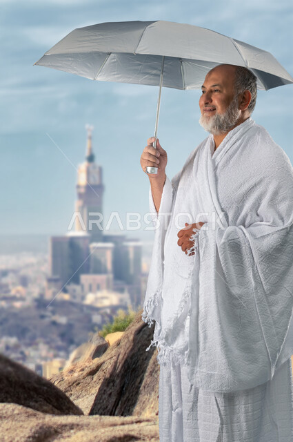  الوقوف على جبل عرفات, رجل مسن حاج يرتدي ثياب الإحرام و يحمل مظلة رمادية, أداء مناسك الحج و العمرة, موسم الحج 2023, ايماءات وجه تدل على السعادة و الخشوع, خلفية برج الساعة في مكة المكرمة.
