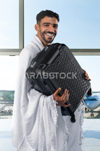 الاستعداد للتوجه و الانطلاق لزيارة بيت الله الحرام, شاب عربي خليجي سعودي يرتدي ملابس الاحرام و يحمل حقيبة سفر سوداء, أداء فريضة الحج و العمرة في موسم الحج 2023, ايماءات وجه تدل على السعادة والحماس للوصول لمكة المكرمة, خلفية  المطار.