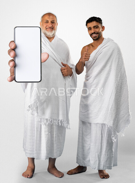 استخدام تطبيقات الهاتف المحمول لارشادات الحج و العمرة, الدليل الالكتروني للحاج و المعتمر, بورتريه لرجلان عربيان خليجيان سعوديان يرتديان ثياب الإحرام, موسم الحج 2023, جوال كبير الحجم بشاشة بيضاء فارغة,  خلفية رمادية.