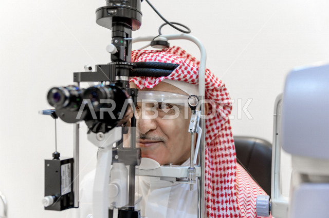 رجل عربي خليجي سعودي في عيادة مختصة لفحص العيون ينظر من خلال مجموعة من العدسات ، طبيبة سعودية خليجية تقوم بمجموعة من الاختبارات لتقييم الرؤية والكشف عن أمراض العيون للمريض ، تقديم الخدمات والرعاية الصحية