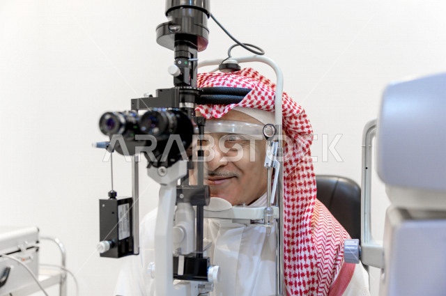 رجل عربي خليجي سعودي في عيادة مختصة لفحص العيون ينظر من خلال مجموعة من العدسات ، طبيبة سعودية خليجية تقوم بمجموعة من الاختبارات لتقييم الرؤية والكشف عن أمراض العيون للمريض ، تقديم الخدمات والرعاية الصحية