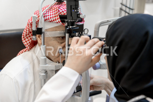 رجل عربي خليجي سعودي في عيادة مختصة لفحص العيون ينظر من خلال مجموعة من العدسات ، طبيبة سعودية خليجية تقوم بمجموعة من الاختبارات لتقييم الرؤية والكشف عن أمراض العيون للمريض ، تقديم الخدمات والرعاية الصحية