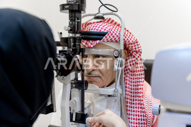 رجل عربي خليجي سعودي في عيادة مختصة لفحص العيون ينظر من خلال مجموعة من العدسات ، طبيبة سعودية خليجية تقوم بمجموعة من الاختبارات لتقييم الرؤية والكشف عن أمراض العيون للمريض ، تقديم الخدمات والرعاية الصحية