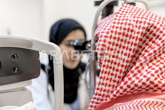 رجل عربي خليجي سعودي في عيادة مختصة لفحص العيون ينظر من خلال مجموعة من العدسات ، طبيبة سعودية خليجية تقوم بمجموعة من الاختبارات لتقييم الرؤية والكشف عن أمراض العيون للمريض ، تقديم الخدمات والرعاية الصحية
