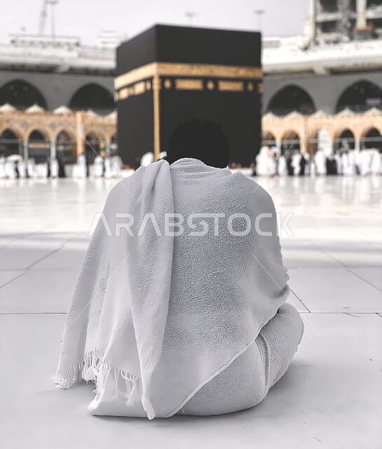A picture from the back of a Saudi Arabian Gulf man sitting in the courtyard of the Holy Mosque of Mecca, wearing the Ihram dress, performing the rituals of Hajj and Umrah, circumambulating around the Kaaba, worshiping and drawing closer to God, Islamic r