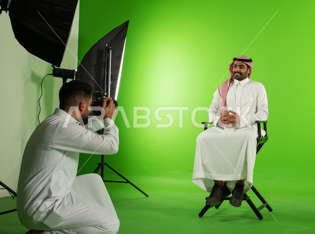 كروما لجلسة تصوير في الاستوديو، خلفية شاشة خضراء، مصور محترف عربي سعودي خليجي يقوم بالتقاط صور فوتوغرافية لرجل يرتدي الزي السعودي التقليدي، كاميرات ومعدات متطورة، تصوير اعلاني تسويقي، خلف الكواليس