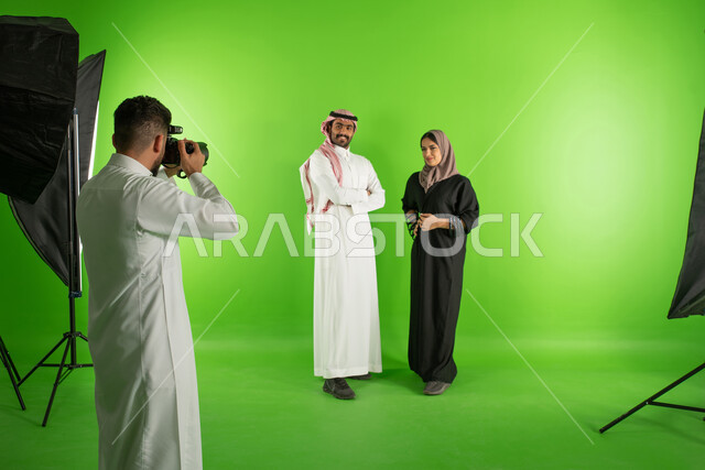 كروما لجلسة تصوير في الاستوديو، مصور محترف عربي سعودي خليجي يقوم بالتقاط صور فوتوغرافية لرجل وامرأة يرتديان الزي السعودي التقليدي، كاميرات ومعدات متطورة، تصوير اعلاني تسويقي، أزياء وموديلات عصرية، خلفية شاشة خضراء، خلف الكواليس